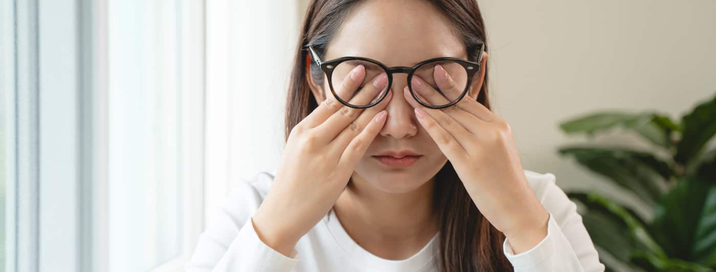 Woman with glasses sits in a room and covers her eyes so people can't see that she is taking drugs that cause glossy eyes.